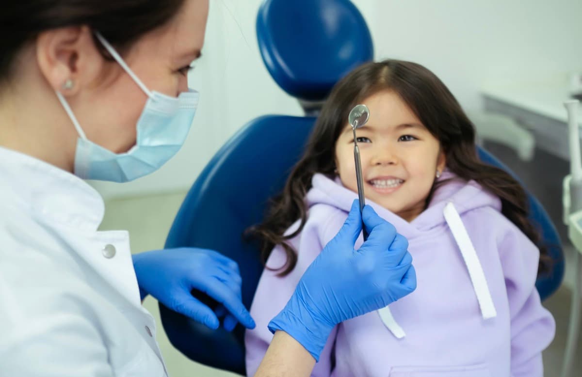 Prima vizită copil dentist Bacău — fetiță zâmbitoare și relaxată în scaunul stomatologic, medic pedodont arată cu oglinda dentară în timpul consultației pediatrice la Chromatic Dental Clinic, atmosferă caldă fără frică